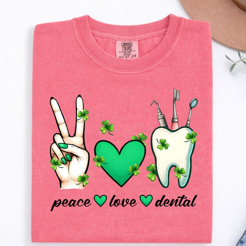 May include: A coral pink t-shirt featuring a graphic with a hand making a peace sign, a green heart, and a white tooth with dental tools, all decorated with green shamrocks. The text below reads peace, love, and dental with small heart icons.