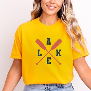 May include: A bright yellow short-sleeved t-shirt featuring a graphic design with two crossed orange oars. The letters L, A, K, and E are printed in a dark navy blue block font, arranged around the oars to spell the word LAKE.