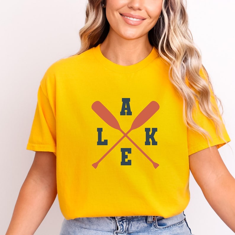 May include: A bright yellow short-sleeved t-shirt featuring a graphic design with two crossed orange oars. The letters L, A, K, and E are printed in a dark navy blue block font, arranged around the oars to spell the word LAKE.
