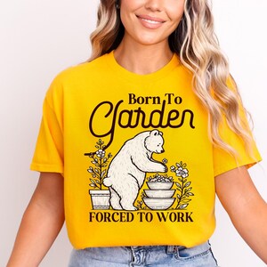 May include: A bright yellow short-sleeved t-shirt featuring a dark brown graphic of a bear standing beside potted flowers and a stack of bowls. The text reads Born To Garden at the top and FORCED TO WORK at the bottom in a bold dark brown font.