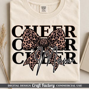 May include: A cream-colored t-shirt featuring the black text CHEER repeated three times in a stacked layout. A large leopard print bow graphic is centered over the text, with the word Mom written in white cursive script over the bow.