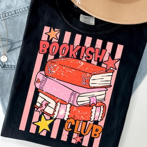 May include: A black t-shirt featuring a graphic print of three stacked books in red and pink with the text "BOOKISH CLUB" in orange. The design includes pink vertical stripes and yellow and red stars, with a distressed texture throughout the print.