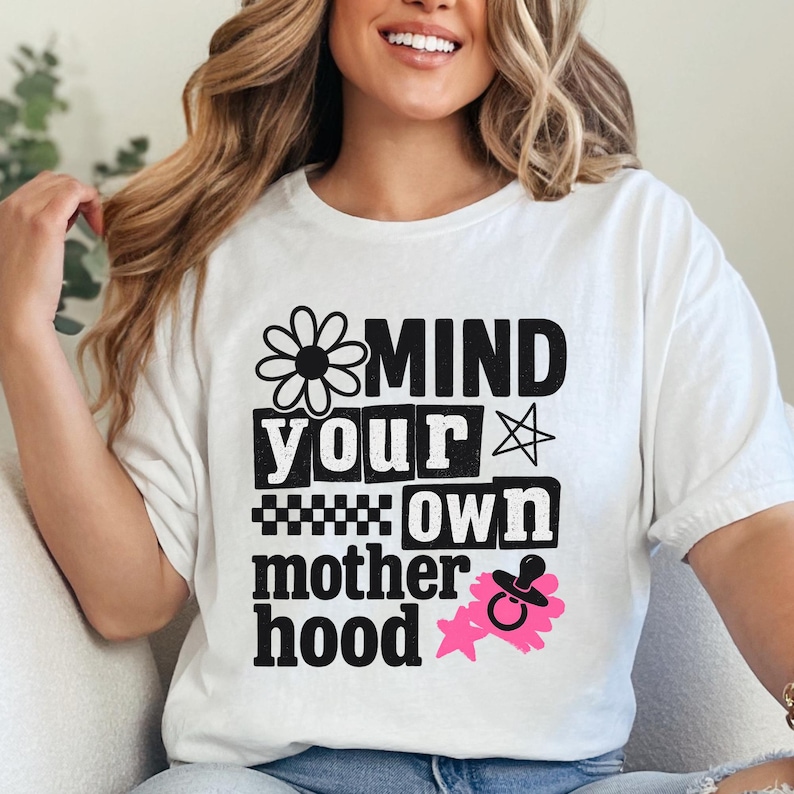 May include: A white short-sleeved t-shirt featuring black text that reads "MIND YOUR OWN motherhood" with a daisy icon, a star, a checkered pattern, and a pink graphic of a baby pacifier with a starburst shape behind it.