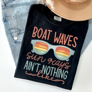 May include: A black t-shirt featuring the text BOAT WAVES in coral, a light blue pair of sunglasses with rainbow-striped lenses, and the text sun rays AIN'T NOTHING like in coral and light blue script and block lettering on the front.