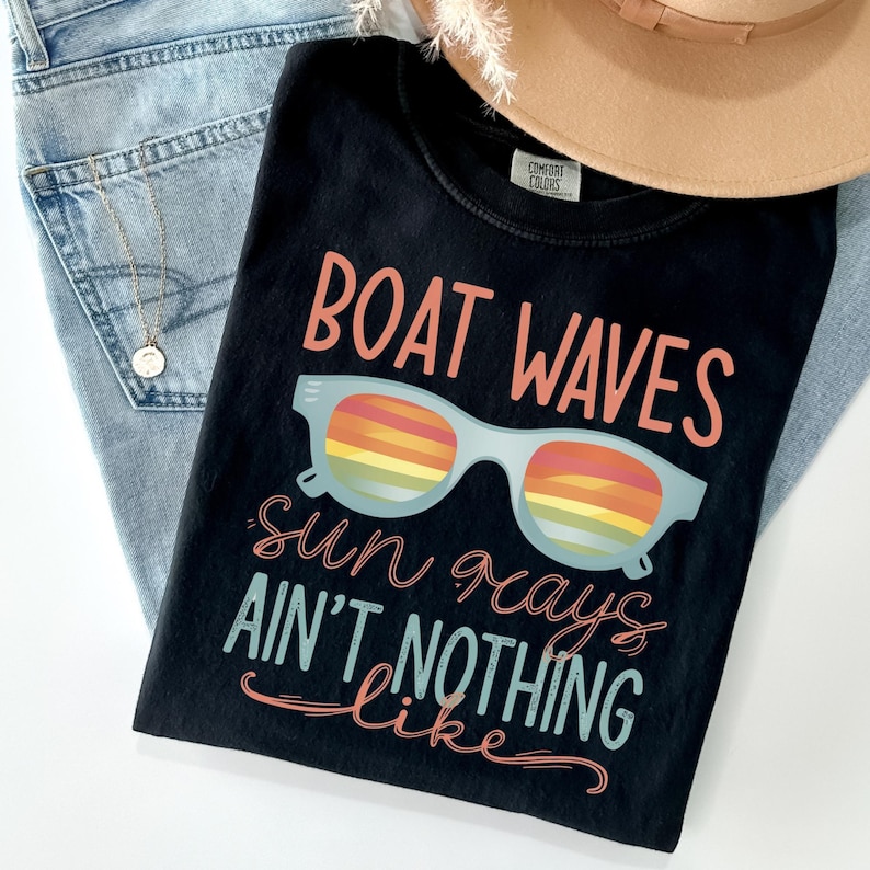 May include: A black t-shirt featuring the text BOAT WAVES in coral, a light blue pair of sunglasses with rainbow-striped lenses, and the text sun rays AIN'T NOTHING like in coral and light blue script and block lettering on the front.