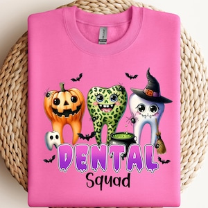 May include: A bright pink crewneck sweatshirt featuring a graphic of three cartoon teeth characters. One tooth is an orange jack-o'-lantern, one has a green leopard print, and one wears a witch hat. The text reads DENTAL Squad in purple.