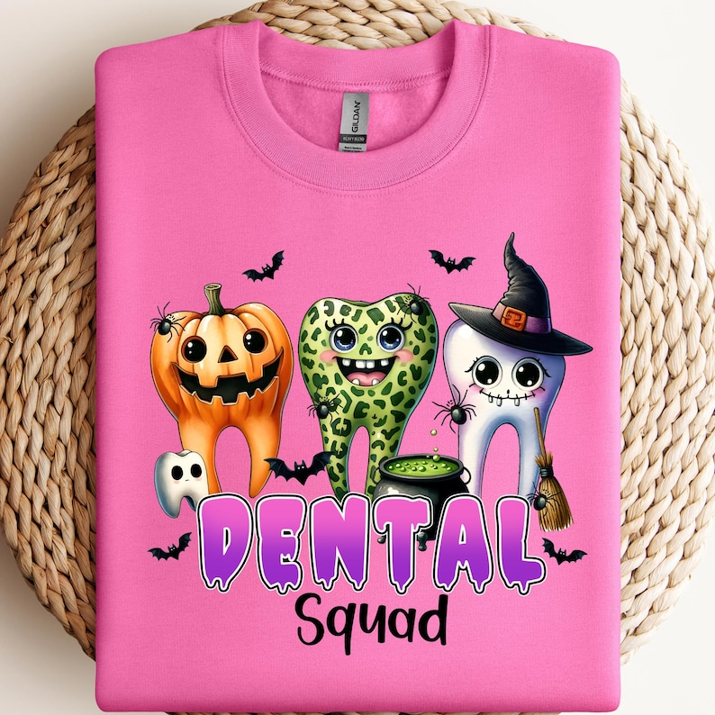 May include: A bright pink crewneck sweatshirt featuring a graphic of three cartoon teeth characters. One tooth is an orange jack-o'-lantern, one has a green leopard print, and one wears a witch hat. The text reads DENTAL Squad in purple.