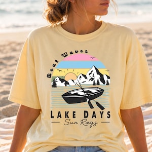 May include: A pale yellow short-sleeve t-shirt featuring a graphic print of a black boat on water with oars, mountains, a sun, and birds. The text reads Boat Waves, LAKE DAYS, and Sun Rays in black lettering against a colorful sunset background.