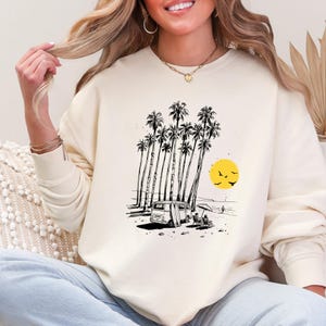 May include: Cream-colored sweatshirt featuring a black and white beach scene. The graphic includes palm trees, a van with a surfboard, a yellow sun, and birds. The sweatshirt is a relaxed fit.