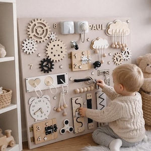 Könnte beinhalten: Ein beiges Holz-Sensorik-Board mit interaktiven Elementen wie Zahnrädern, Riegeln, Schaltern und einer Uhr. Der Name "PAUL" steht oben. Ein Kind interagiert mit dem Board auf einem weißen, flauschigen Teppich. Das Board ist für Kleinkinder.