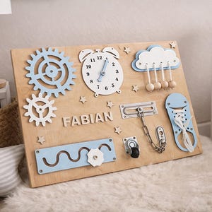 May include: Wooden activity board with various interactive elements. Features a clock, gears, a cloud with hanging beads, and a shoe with laces. Includes the name "FABIAN" in wooden letters. The board is light brown with blue and white accents.