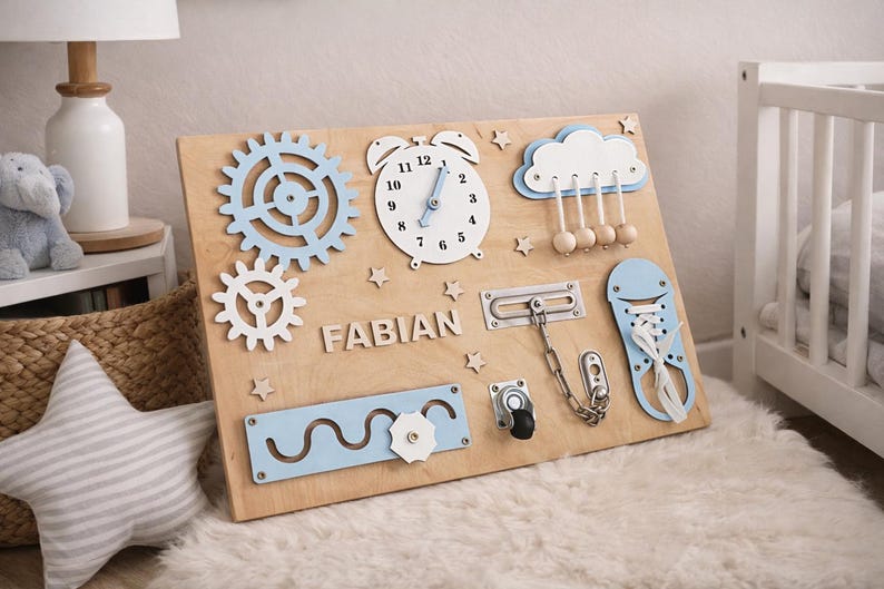 May include: Wooden activity board with various interactive elements. Features a clock, gears, a cloud with hanging beads, and a shoe with laces. Includes the name "FABIAN" in wooden letters. The board is light brown with blue and white accents.