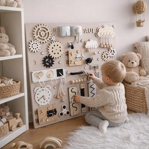 May include: A beige wooden activity board with various interactive elements, including gears, latches, and a clock. The board has the name "PAUL" at the top. A child is interacting with the board on a white fluffy rug.