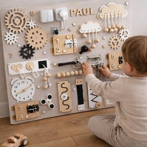 May include: A beige wooden activity board with various interactive elements, including gears, latches, switches, and a clock. The board features the name "PAUL" and is designed for child development and sensory exploration. A child is interacting with the board.