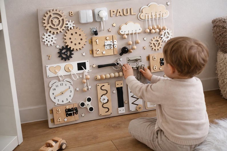 May include: A beige wooden activity board with various interactive elements, including gears, latches, switches, and a clock. The board features the name "PAUL" and is designed for child development and sensory exploration. A child is interacting with the board.