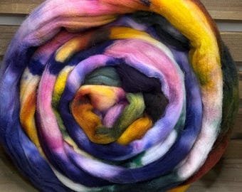 May include: A thick, coiled bundle of unspun wool roving featuring a vibrant, multi-colored tie-dye pattern. The fibers display a swirling blend of bright yellow, deep purple, magenta, orange, and dark green hues, creating a soft, textured appearance.
