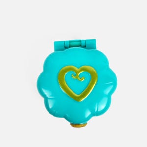 Op de afbeelding: Een turquoise bloemvormig speelgoed met een gouden hartontwerp in het midden. Het speelgoed heeft een scharnierend deksel en een kleine, goudkleurige basis. Het speelgoed is een vintage verzamelobject.