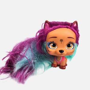 Peut inclure: Un chiot jouet marron et rose avec de grands yeux expressifs et de longs cheveux colorés dans les tons violets et turquoise. Le chiot porte un collier rose et a un motif floral sur le visage. Le jouet est conçu pour ressembler à un chiot.