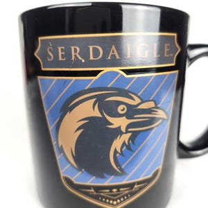 Può includere: Tazza in ceramica nera con la scritta "SERDAIGLE" in oro. La tazza presenta uno stemma con un disegno di corvo in oro e nero, su sfondo blu a strisce. La tazza ha un manico nero.