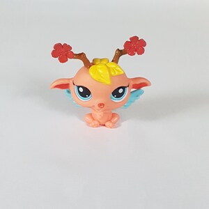 Pode incluir: Uma pequena figura de fada rosa com asas azuis, cabelo de folha amarela e chifres de flores vermelhas.