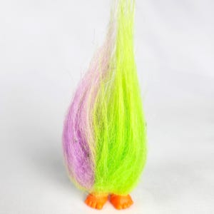 Puede incluir: Una muñeca troll vintage con pelo difuso verde neón y morado. La muñeca tiene pies naranjas y un cuerpo redondo. El pelo es largo y se levanta. Es un juguete clásico.