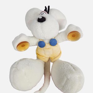 Könnte beinhalten: Ein weißes Plüschmaus-Spielzeug mit großen Ohren, schwarzen Augen und einer rosa Nase. Die Maus trägt eine gelbe Latzhose mit blauen Knöpfen. Die Hände und Füße haben gelbe Akzente. Das Spielzeug hat einen langen, dünnen Schwanz.