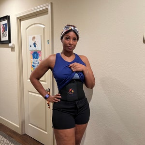May include: A woman wearing a blue tank top, black shorts, and a black waist trainer with a red and orange logo. She is wearing a patterned headband and a smartwatch on her left wrist.