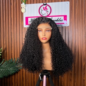 May include: A black curly wig on a mannequin head, displayed on a pink tripod stand. The wig has a lace front and is styled with voluminous, tight curls. A sign in the background reads "Your Sparkles Beauty".