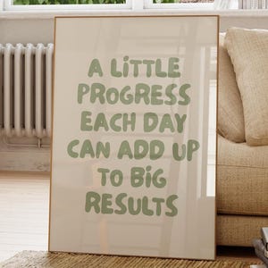 Könnte beinhalten: Ein gerahmter Druck mit dem Text "A Little Progress Each Day Can Add Up To Big Results" in grünen Buchstaben. Der Druck lehnt an einem beigen Sofa und einem Stapel Bücher. Eine Topfpflanze und ein Fenster sind im Hintergrund.