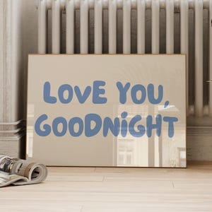 May include: A rectangular wall art print featuring a light beige background with the text "LOVE YOU, GOODNIGHT" written in a bold, rounded, periwinkle blue font. The print is displayed in a simple frame, showcasing clear, legible lettering.