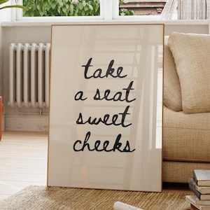 Puede incluir: Un cuadro enmarcado con las palabras "take a seat sweet cheeks" en escritura negra. El cuadro está en una habitación con un sofá beige, una planta en maceta y una ventana con vistas a un edificio.
