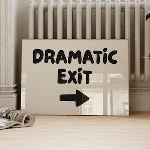 May include: A framed sign with the words "DRAMATIC EXIT" in bold, black letters and a black arrow pointing right. The sign is beige and rests on a light wood floor. A radiator and a potted plant are in the background.
