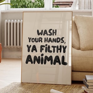 Può includere: Una stampa incorniciata con la scritta "WASH YOUR HANDS, YA FILTHY ANIMAL" in grassetto nero. La stampa ha uno sfondo beige e una cornice dorata. La stampa è esposta in una stanza ben illuminata con una finestra e un divano beige.