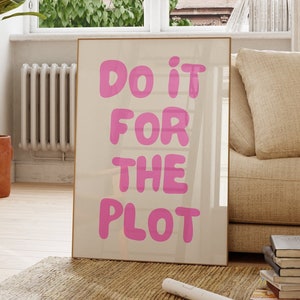 May include: A framed poster with the text "Do it for the plot" in pink bubble letters on a white background. The poster is leaning against a beige couch in a room with a large window and a potted plant.