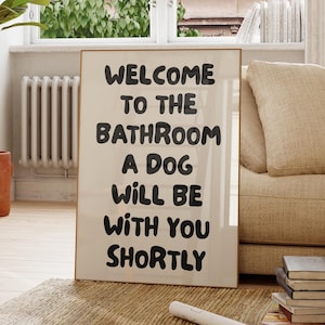 May include: A framed sign with the text "WELCOME TO THE BATHROOM A DOG WILL BE WITH YOU SHORTLY" in black lettering. The sign is in a room with a window, a plant, and a beige sofa.