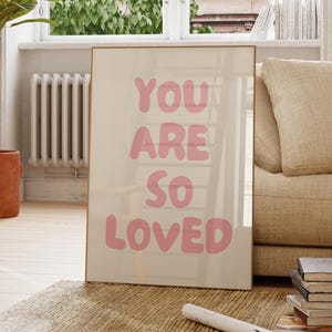 以下が含まれることがあります： ピンク色の文字で「YOU ARE SO LOVED」と書かれたフレーム入りのプリント。ベージュのソファ、木製の椅子、鉢植えのある部屋の壁に立てかけられています。ラグと窓も写っています。