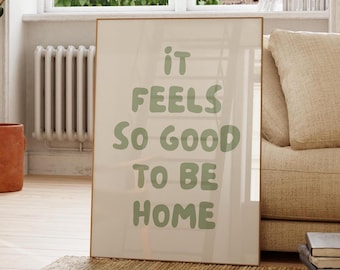 Könnte beinhalten: Gerahmtes Poster mit dem Text "IT FEELS SO GOOD TO BE HOME" in hellgrünen Buchstaben. Das Poster lehnt an einem beigen Sofa auf einem gewebten Teppich. Eine Topfpflanze und ein Holzstuhl befinden sich ebenfalls im Raum.