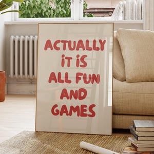 May include: A framed print with the text "ACTUALLY IT IS ALL FUN AND GAMES" in red letters. The print is leaning against a beige sofa in a well-lit room with a large window, a wooden chair, and a potted plant.