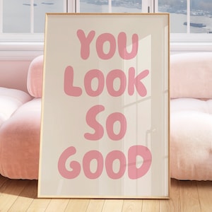 May include: A framed poster with the text "YOU LOOK SO GOOD" in pink letters on a white background. The poster is leaning against a pink sofa in a room with a window.