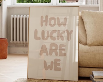 May include: A framed print with the text "HOW LUCKY ARE WE" in a soft, rounded font. The print is leaning against a wall in a room with a large window, a sofa, and a wooden chair. A potted plant and a rug are also visible.