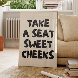 Puede incluir: Un sofá beige con un cojín se encuentra en una sala de estar con una gran impresión enmarcada que dice "Take a seat sweet cheeks". También hay una silla de madera y una planta en maceta en la habitación.