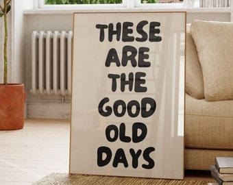 May include: A framed print with the text "These are the good old days" in black lettering on a white background. The print is hanging on a white wall in a room with a large window, a potted plant, and a couch.