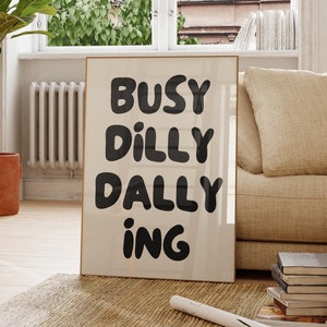 May include: A framed print with the text "Busy Dilly Dallying" in black lettering on a white background. The print is hanging on a white wall in a living room setting with a beige couch, a wooden side table, and a potted plant.