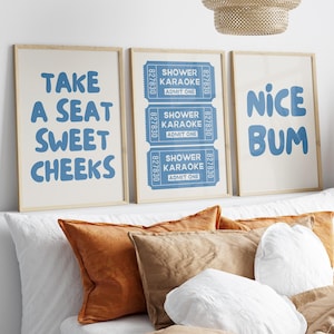 Könnte beinhalten: Drei gerahmte Kunstdrucke mit hellen Holzrahmen. Die Drucke zeigen blauen Text auf cremefarbenem Hintergrund. Der Text enthält Sätze wie "Take a seat sweet cheeks", "Shower Karaoke" und "Nice Bum". Die Drucke werden über einem Bett mit orangefarbenen und weißen Kissen angezeigt.