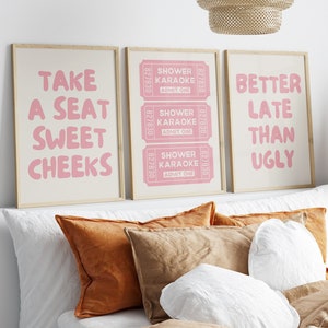May include: Three framed prints with pink text on a white background. The prints are hanging on a white wall above a bed with brown and white pillows. The text on the prints reads "Take a seat sweet cheeks", "Shower Karaoke Admit One" and "Better Late Than Ugly".