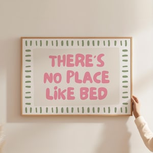 Puede incluir: Impresión enmarcada con el texto "THERE'S NO PLACE LIKE BED" en letras rosas. La impresión tiene un fondo beige claro con un borde blanco y guiones verdes. El marco es de color marrón claro.
