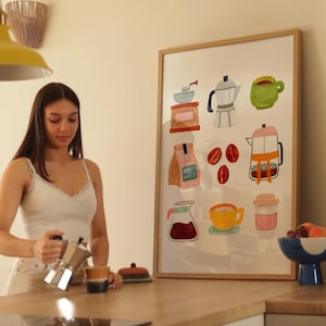 May include: A framed watercolor illustration of coffee making tools and ingredients. The illustration features a coffee grinder, coffee beans, a coffee pot, a mug, a measuring cup, and a bag of coffee. The illustration is in a light pink, yellow, green, and brown color palette.