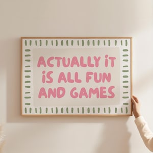 May include: A rectangular wall art print in a light wood frame featuring the pink text "ACTUALLY IT IS ALL FUN AND GAMES" centered on a beige background. The print is bordered by a repeating pattern of short, vertical sage green dash marks.