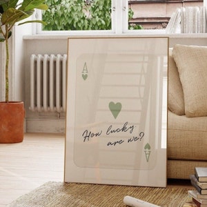 Puede incluir: Una impresi&oacute;n enmarcada con fondo blanco y un dise&ntilde;o de carta de juego de as de corazones verde. El texto "How lucky are we?" est&aacute; escrito en cursiva negra.
