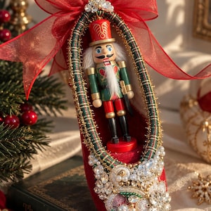 May include: A red velvet shoe ornament featuring a central nutcracker figure in a green and red uniform. The shoe is trimmed with green plaid fabric, gold metallic fringe, and an elaborate cluster of clear crystals and white pearls at the toe.